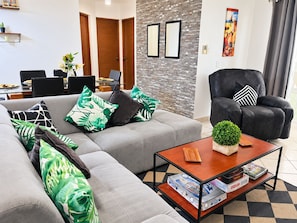 Una sala de estar moderna con un sofá gris, un sillón negro y una mesa de centro. Hay varios cojines estampados en verde y negro sobre el sofá.
