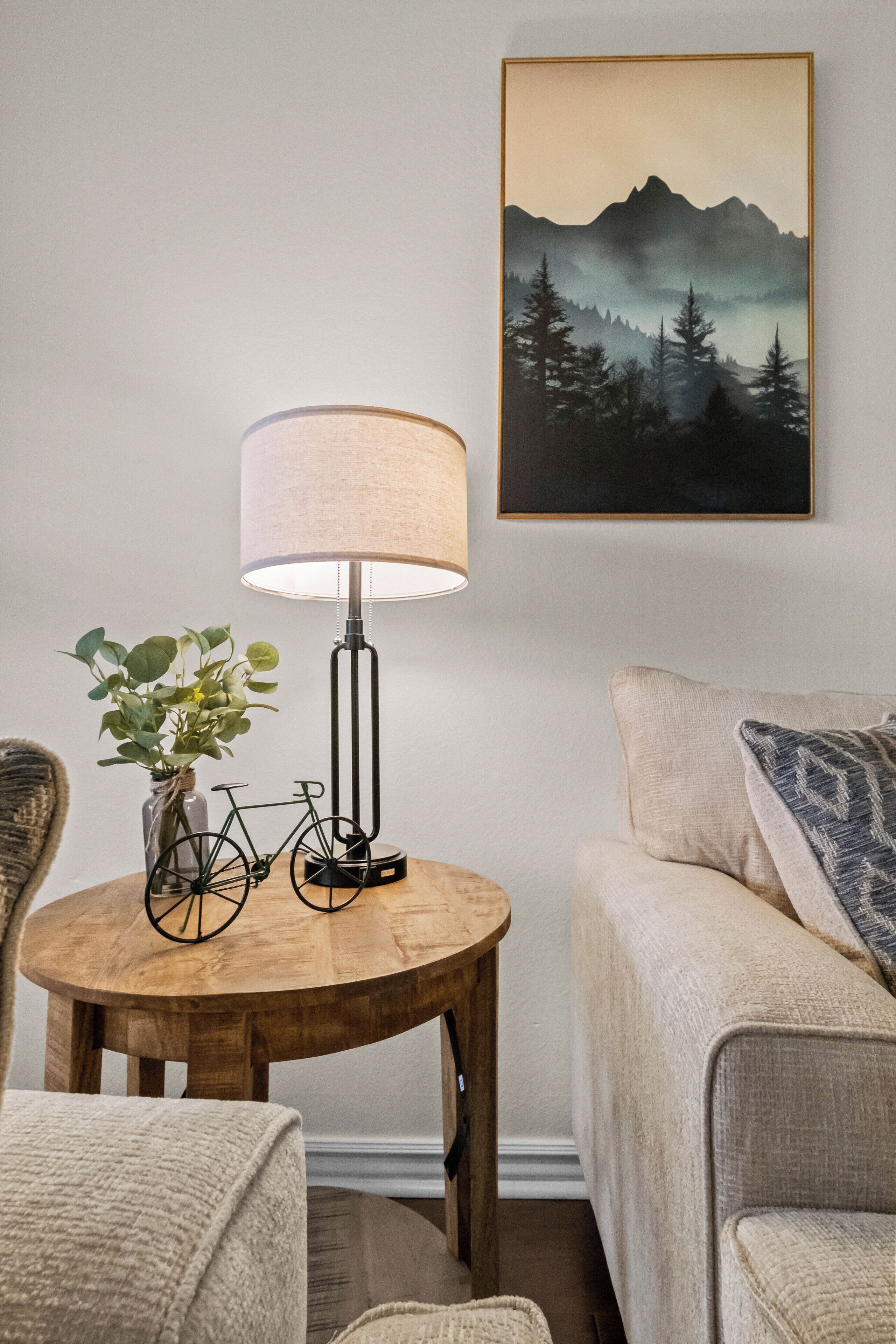 A cozy room with a wooden table, a lamp, a framed landscape painting, and a sofa.