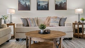 A living room with a beige sofa, patterned cushions, a round wooden coffee table, and three framed landscape paintings on the wall. - Greenway Getaway: Central to NWA & on the Razorback Greenway (Rogers)
