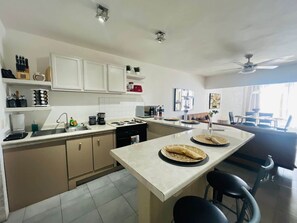 Une cuisine moderne avec des meubles blancs, un îlot central, un coin repas avec un canapé et un ventilateur de plafond.