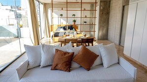 A modern living room with a white sofa, brown cushions, a dining area with wooden furniture, and a view of buildings through large windows.
