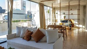 A modern living room with a white sofa, a dining area, and large windows offering a city view.