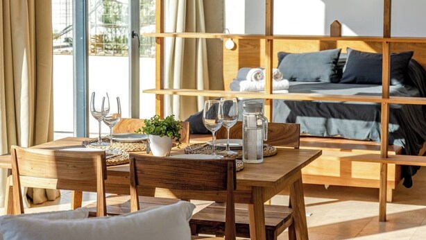 A wooden dining table with a plant, wine glasses, and a water bottle. A bed with pillows and a headboard is visible in the background.