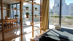 A modern room with a large window, a wooden shelf with potted plants, a dining area with a table and chairs, and a bed with a black cover.