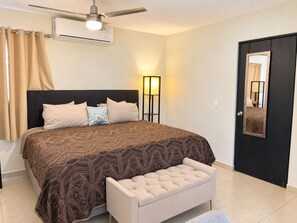Un dormitorio con cama, mesita de noche, banca, ventilador de techo, equipo de aire acondicionado, lámpara de pie y una puerta con espejo.