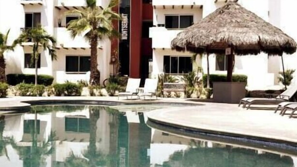 A swimming pool with lounge chairs and a thatched umbrella, surrounded by palm trees and a building with multiple windows. - Chic Cabo Gateaway Walkable to Beach & Downtown Fun (Cabo San Lucas)