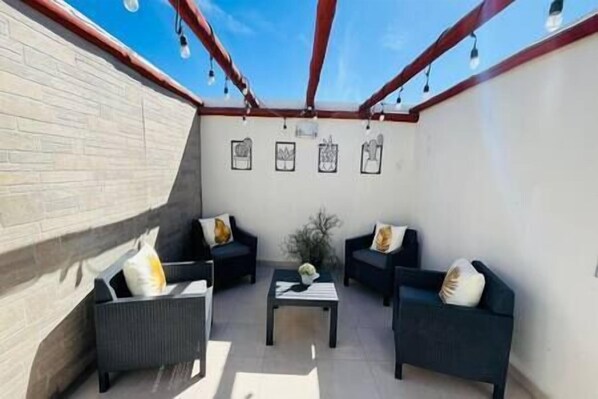 A covered patio area with two sofas and a small table, framed artwork on the wall, and a brick wall.