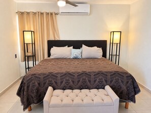 A bedroom with a bed, bedside lamps, an air conditioner, a curtain, and a bench. - Chic Cabo Gateaway Walkable to Beach & Downtown Fun (Cabo San Lucas)