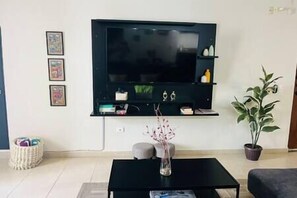 A living room with a flat-screen TV mounted on the wall, a black coffee table, a vase with flowers, a potted plant, and a small side table with decorative items.