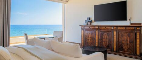 Un dormitorio con un ventanal grande que da al mar, una cama con ropa blanca, un aparador de madera y un televisor de pantalla plana en la pared.