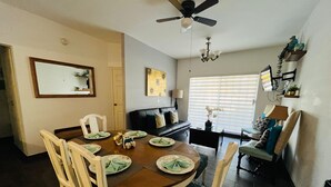 Comedor con una mesa de madera dispuesta para cuatro personas, rodeada de sillas blancas. Se observa un ventilador de techo y una lámpara de araña.