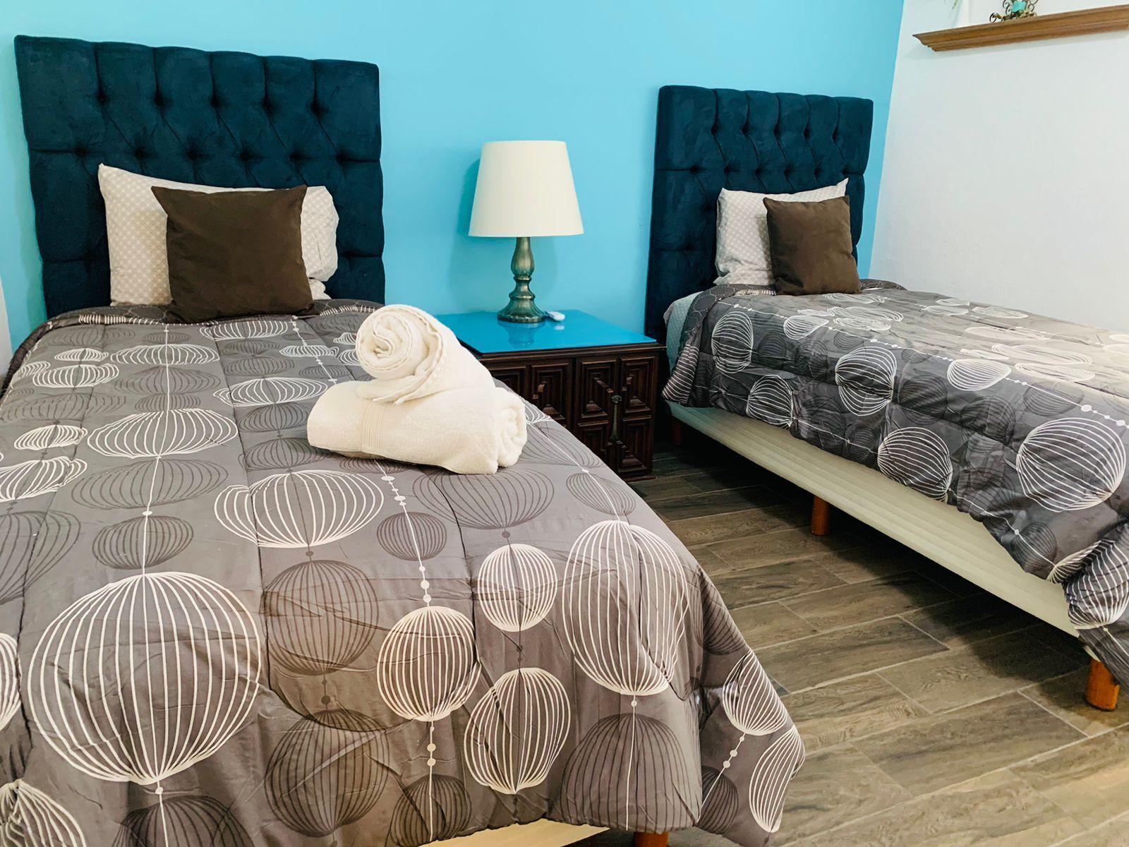 Une chambre d’hôtel avec deux lits, chacun doté d’une tête de lit bleu foncé et d’une couvre-lit à motifs. Une serviette blanche pliée est posée sur le lit, au premier plan. Une petite table avec une lampe se trouve entre les deux lits.