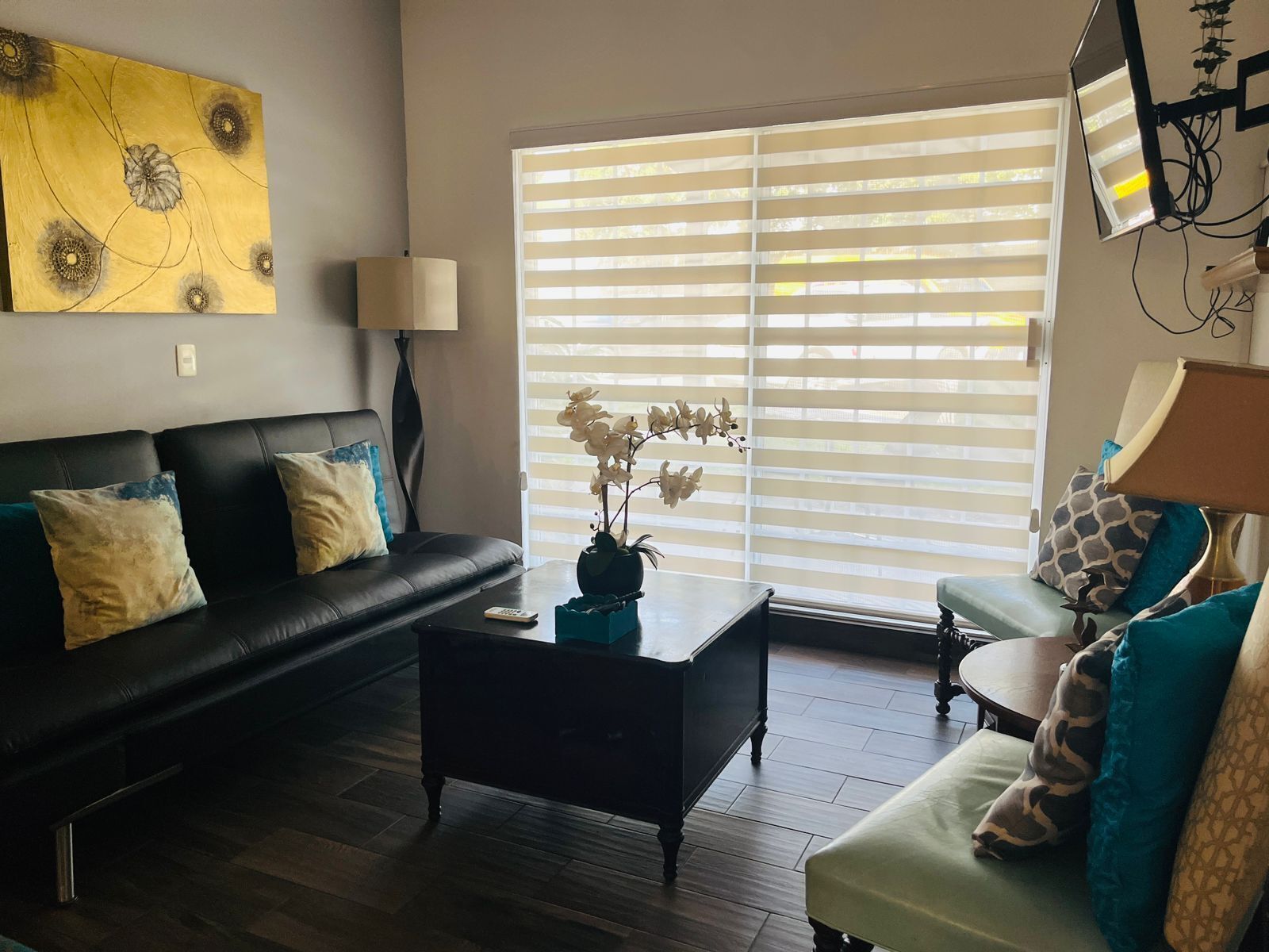 A living room with a black leather sofa, a coffee table, and a green armchair. There is a painting on the wall and a window with blinds.