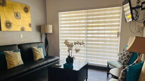 Una sala de estar con un sofá de cuero negro, una mesa de centro y un sillón verde. Hay un cuadro en la pared y una ventana con persianas.