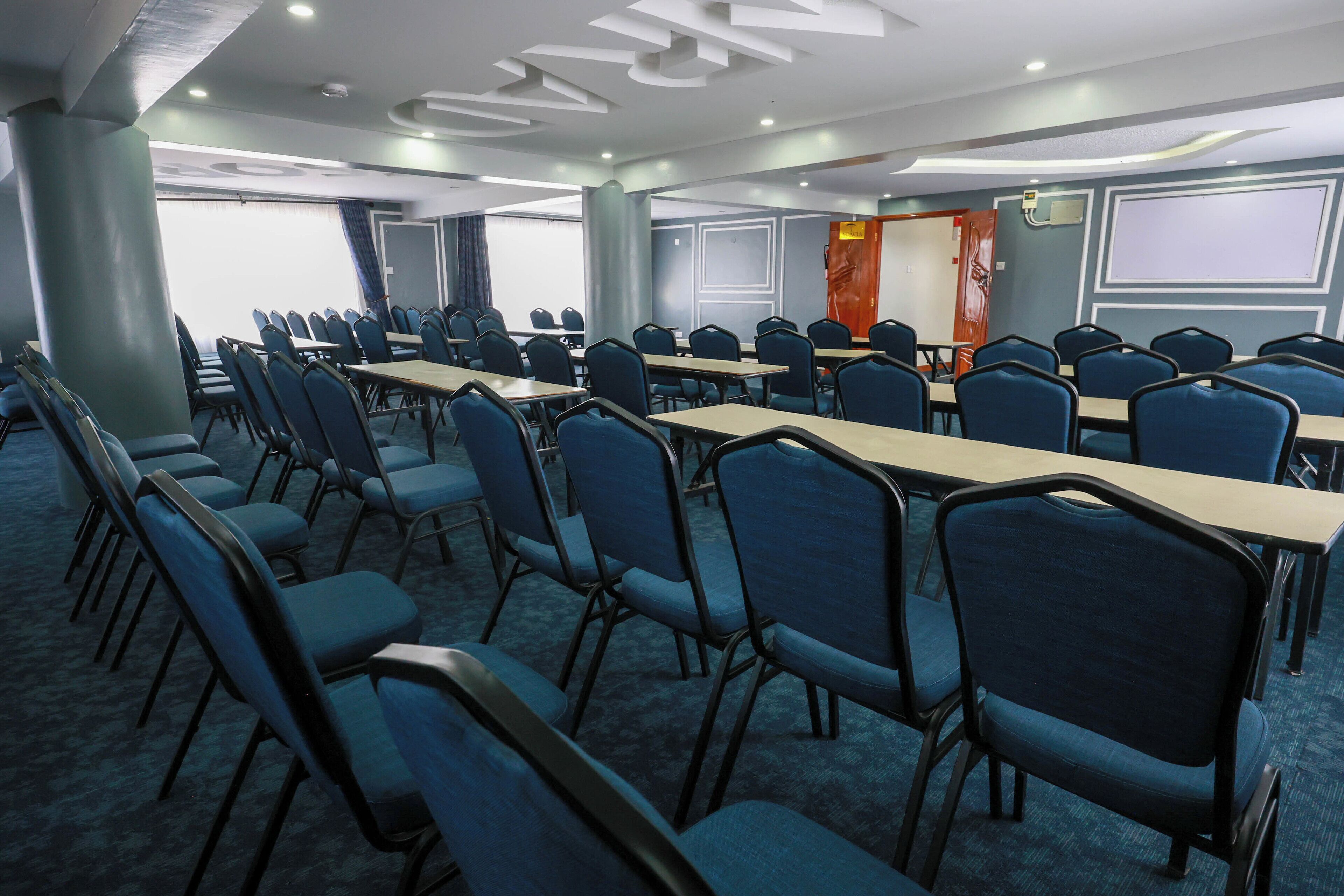 Ein Konferenzraum mit blauen Stühlen und Tischen, einer großen Leinwand und einer Tür mit rotem Griff.