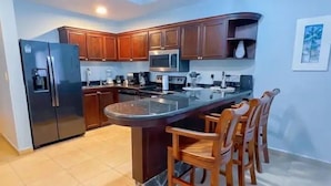 A kitchen with wooden cabinets, a stainless steel refrigerator, a microwave, and a bar area with chairs. - Bahia Encantada G5 (Jacó)