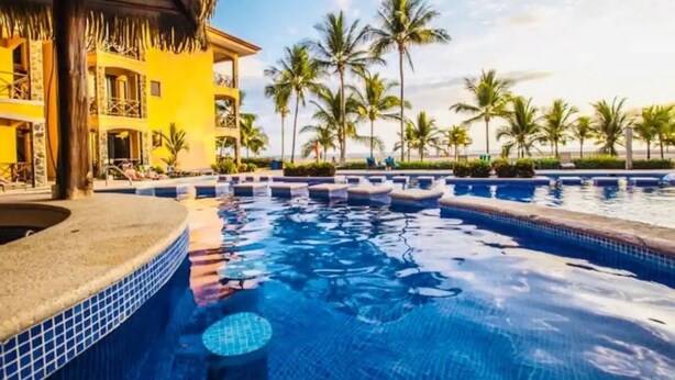 A resort pool area with multiple swimming pools, palm trees, and a building with balconies. - Bahia Encantada G5 (Jacó)