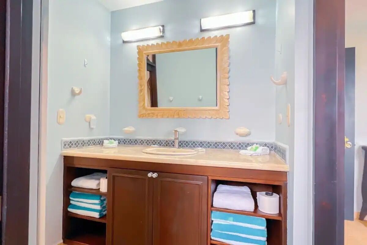 A bathroom with a wooden vanity, a mirror with a scalloped frame, and shelves with folded towels.