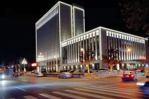 Um edifício moderno com uma estrutura vertical marcante e fachada plana, localizado em um canto de rua com faixas de pedestres e semáforos.