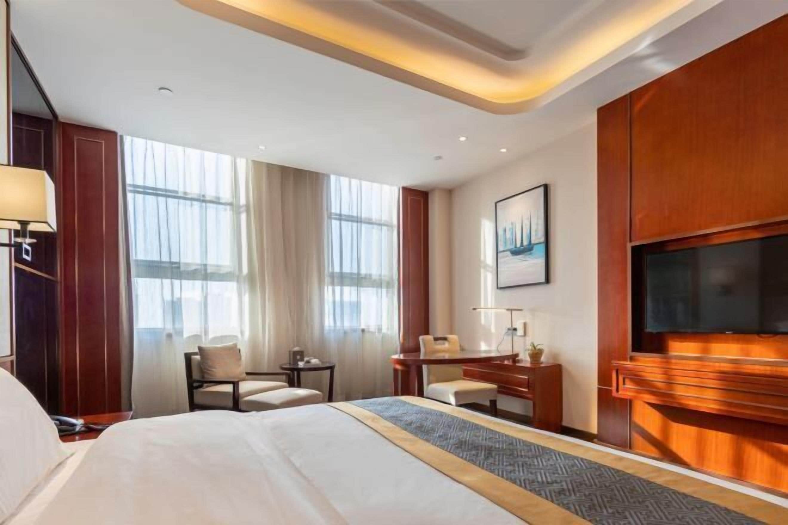A hotel room with a large bed, a desk, a chair, a TV, and a window with curtains.