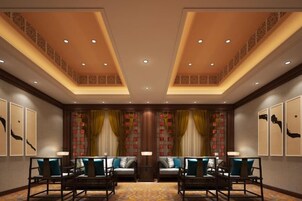 Un vestíbulo de hotel con techo alto, iluminación empotrada, una zona de estar con sillas y sofá, y una pared decorativa con obras de arte enmarcadas.