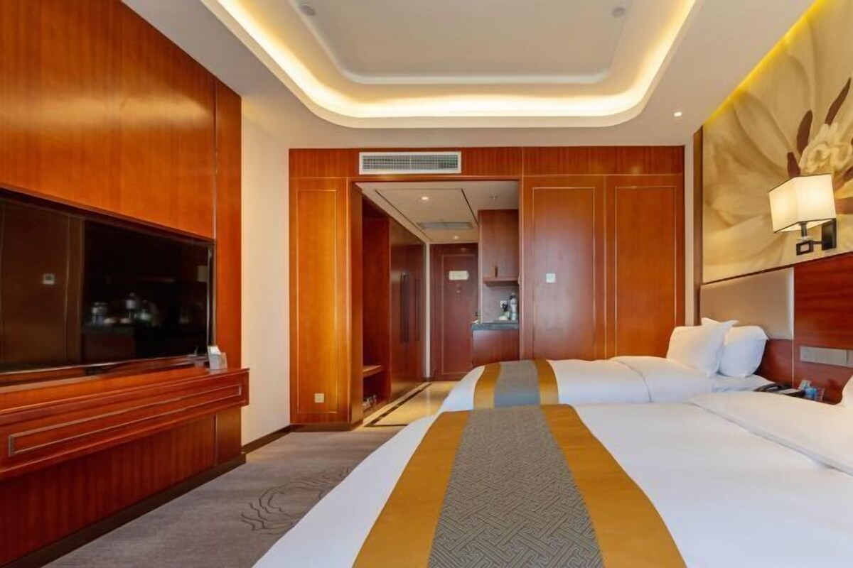 A hotel room with a large TV, a bed with a patterned bedspread, wooden paneling, and a ceiling with recessed lighting.