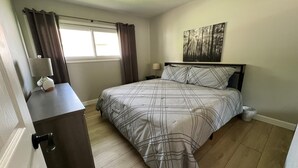 Een slaapkamer met een bed, nachtkastje, lamp, raam met gordijnen en een schilderij aan de muur.