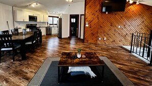 A spacious living area with a wooden herringbone wall, a dining table with chairs, a kitchen with white cabinets, and a ceiling fan.