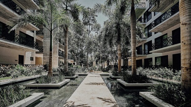 A modern hotel with a central water feature, surrounded by palm trees and concrete planters. - Tulum Stylish 2BR Penthouse W/private Terrace, Grill & Pool (Tulum)
