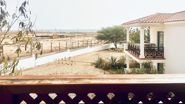 Blick vom Balkon auf einen Sandstrand mit einem weißen Gebäude und einem Baum.
