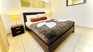 Ein ordentlich bezogenes Bett mit einer grauen Bettdecke, zwei Kissen und zwei roten Zierkissen. Es gibt zwei Nachttische mit Lampen, einen an der Wand befestigten Spiegel und drei gerahmte Kunstwerke über dem Bett.