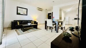 Ein modernes Wohnzimmer mit einer schwarzen Couch, einem Couchtisch und einem Essbereich mit Tisch und Stühlen. Im Hintergrund ist eine Küche erkennbar.
