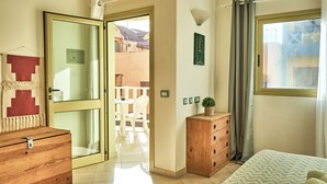 Ein Schlafzimmer mit einem Bett, einer Holzkommode, einem Nachttisch, einem Fenster mit Vorhängen und einer Tür zu einem Außenbereich.