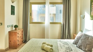Ein Schlafzimmer mit Bett, Kommode, einem Fenster mit Vorhängen und einer Wandleuchte.