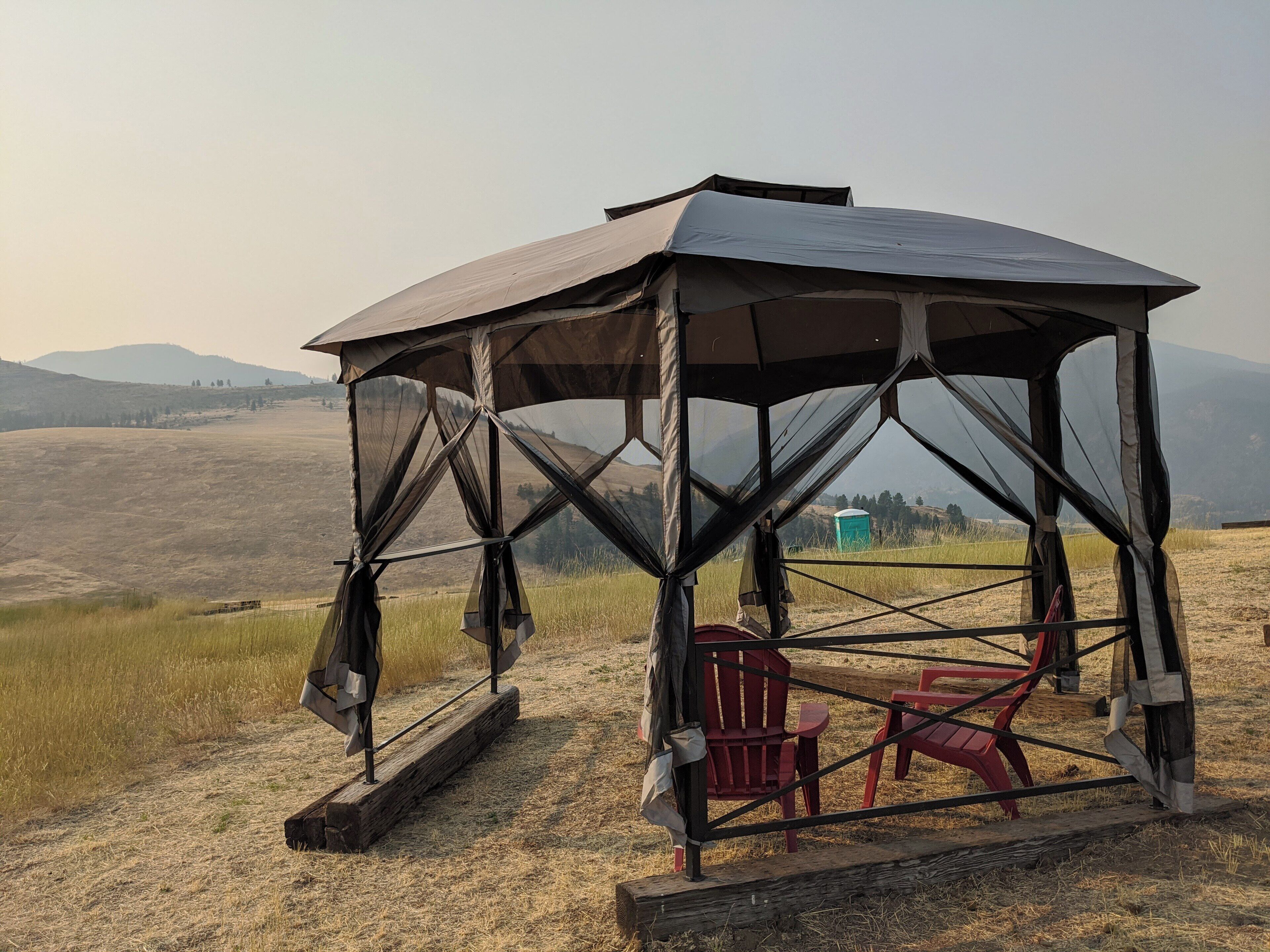 A covered outdoor seating area with a bench and a chair, surrounded by a natural landscape with hills in the background.