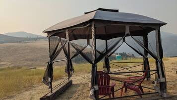 A covered outdoor seating area with a bench and a chair, surrounded by a natural landscape with hills in the background.