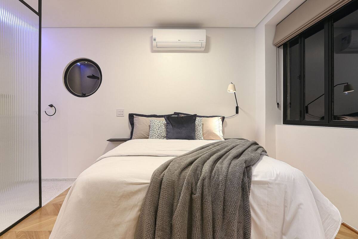 A bedroom with a bed, an air conditioner, a round mirror, a bedside lamp, and a window.