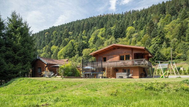 A wooden cabin with a balcony, surrounded by a lush green landscape and a forested mountain backdrop.