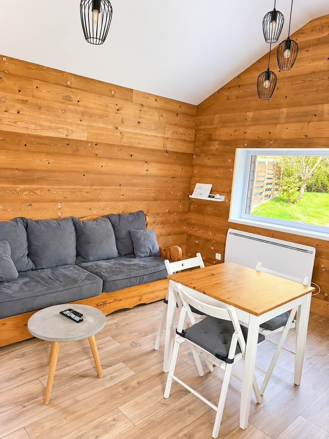 Une pièce chaleureuse avec un décor intérieur en bois, un canapé gris, une table à manger en bois avec des chaises, et une fenêtre donnant sur un paysage verdoyant.