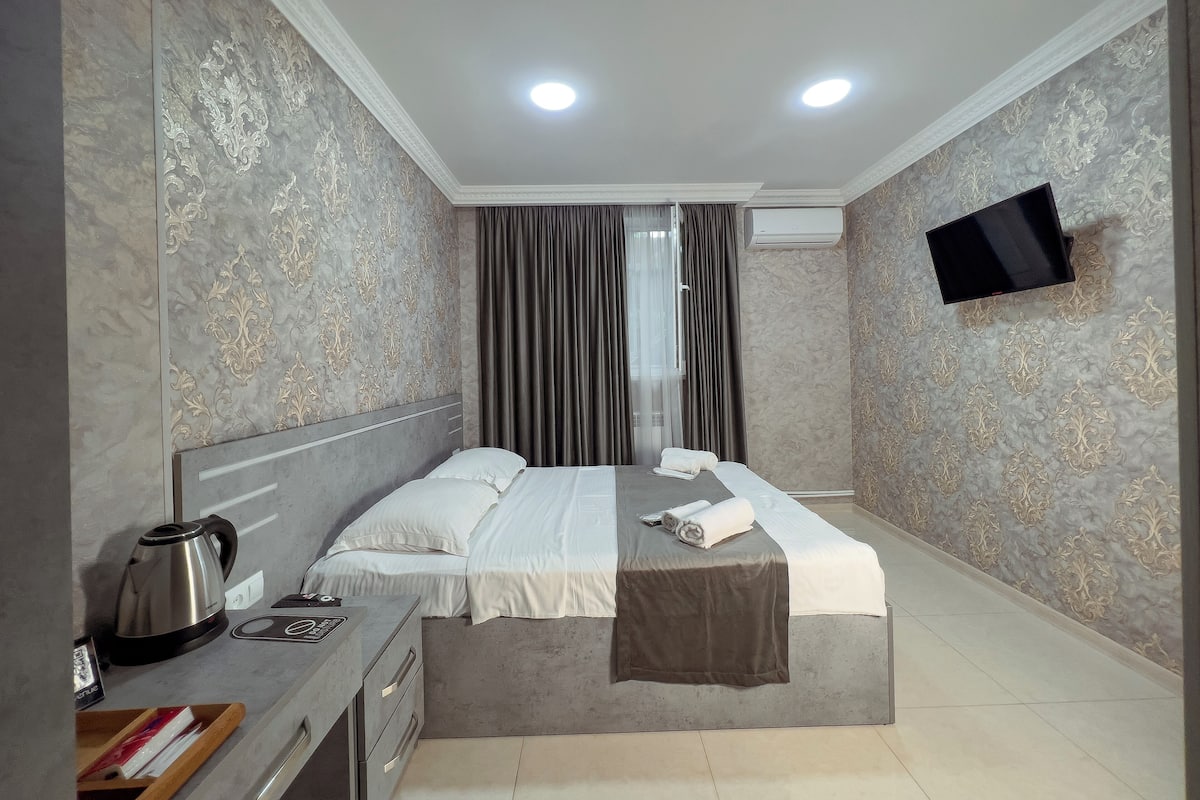 A hotel room with a bed, a desk with a kettle, a flat-screen TV mounted on the wall, and a window with curtains.