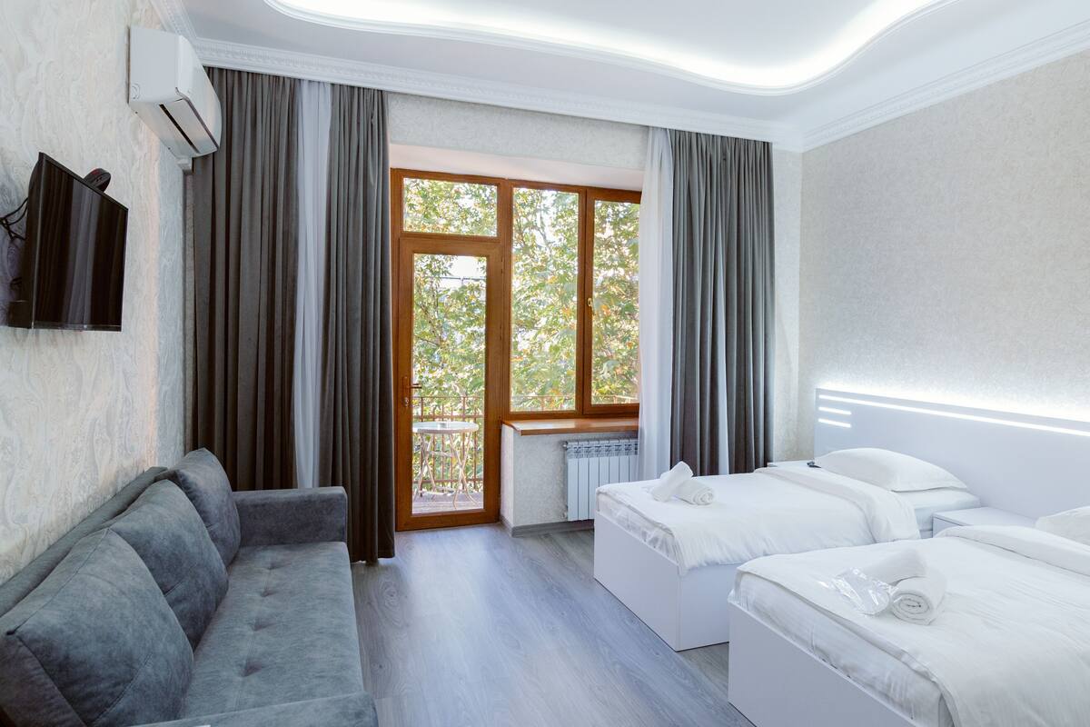A hotel room with a grey sofa, two single beds, a TV mounted on the wall, and a large window with curtains.