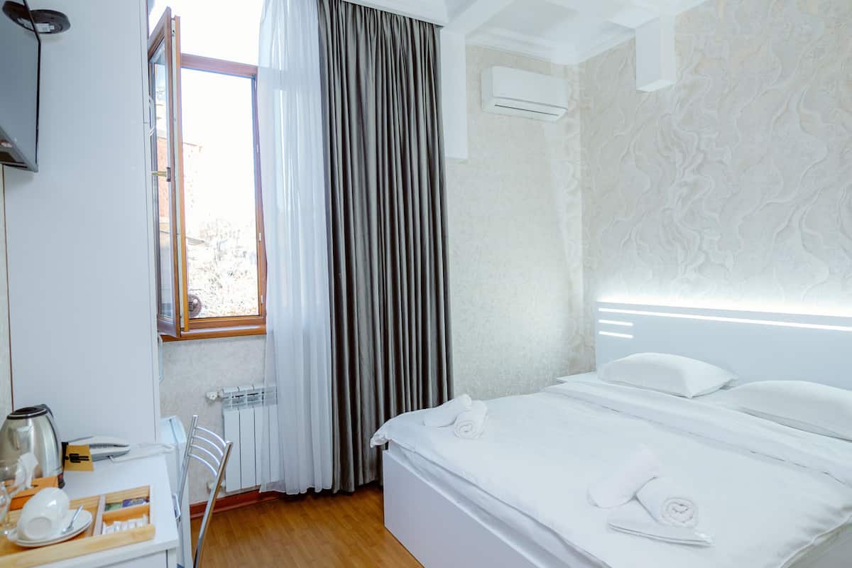 A hotel room with a bed, a desk with a kettle, a chair, and a window with curtains.