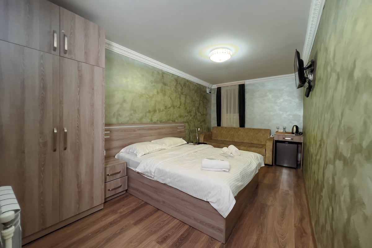 A bedroom with a bed, a wardrobe, a bedside table, a nightstand, a sofa, and a small table with a kettle.
