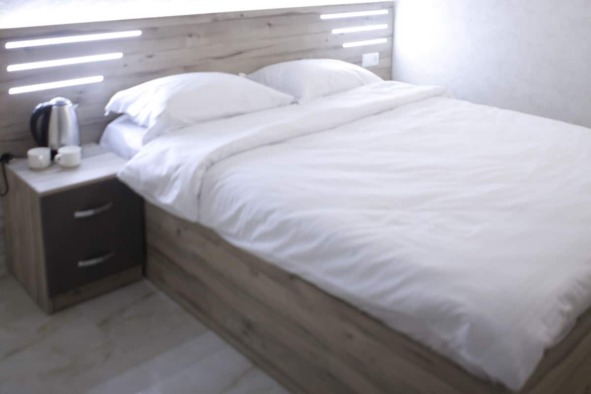 A modern bedroom with a wooden headboard, a bedside table with a kettle, and a white bedspread.