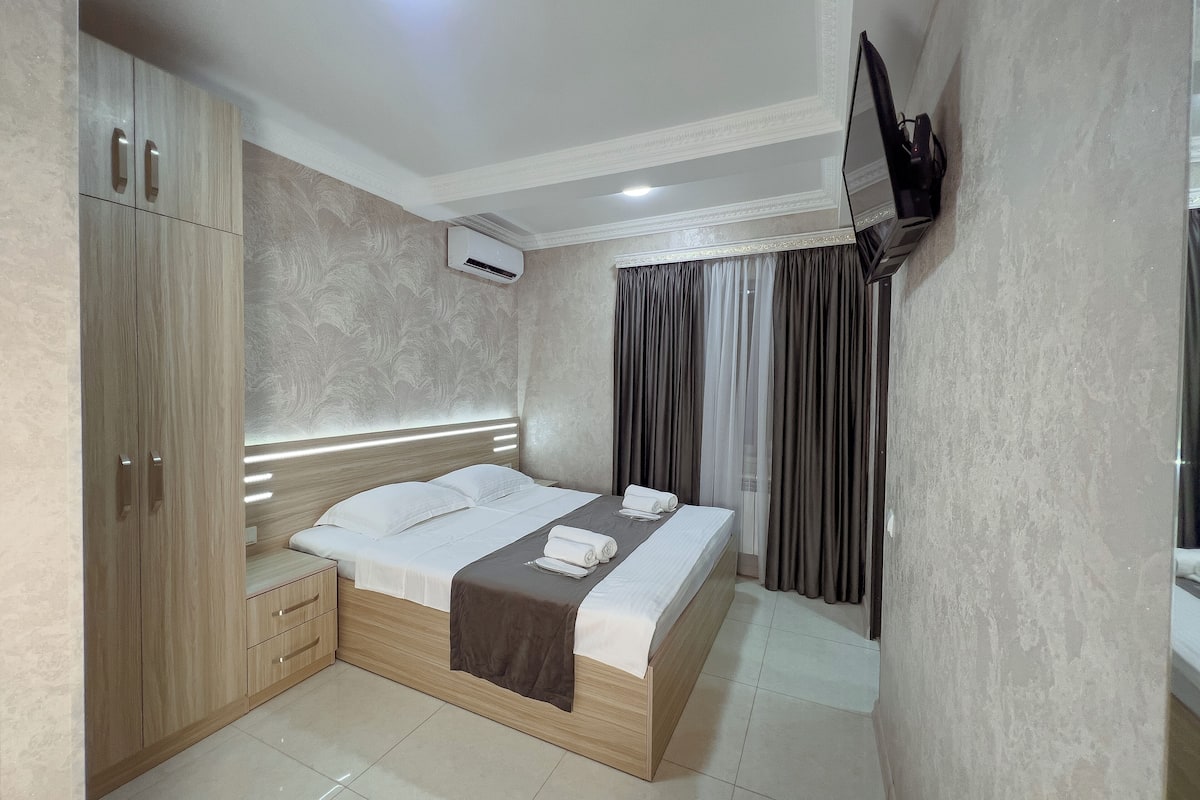A modern hotel room with a bed, wooden wardrobe, and a flat-screen TV mounted on the wall.