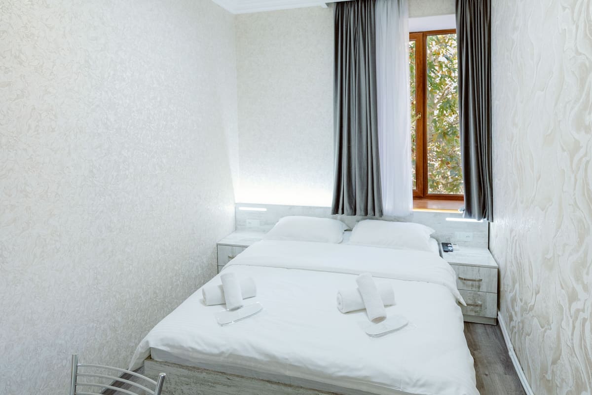 A hotel room with a bed, two bedside tables, a window with curtains, and a view of greenery outside.