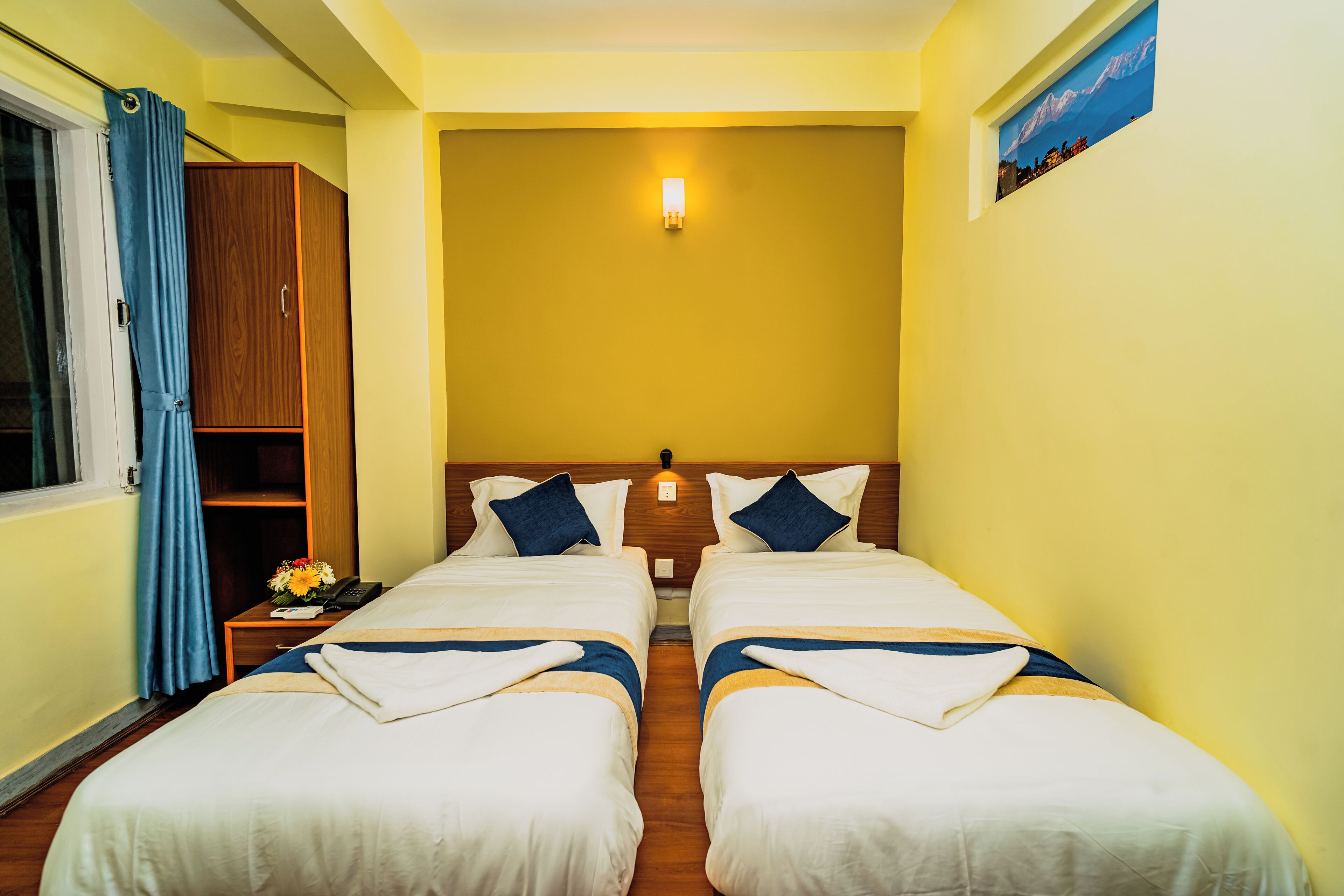 A hotel room with two beds, a wooden wardrobe, a nightstand with a phone and flowers, and a painting on the wall.