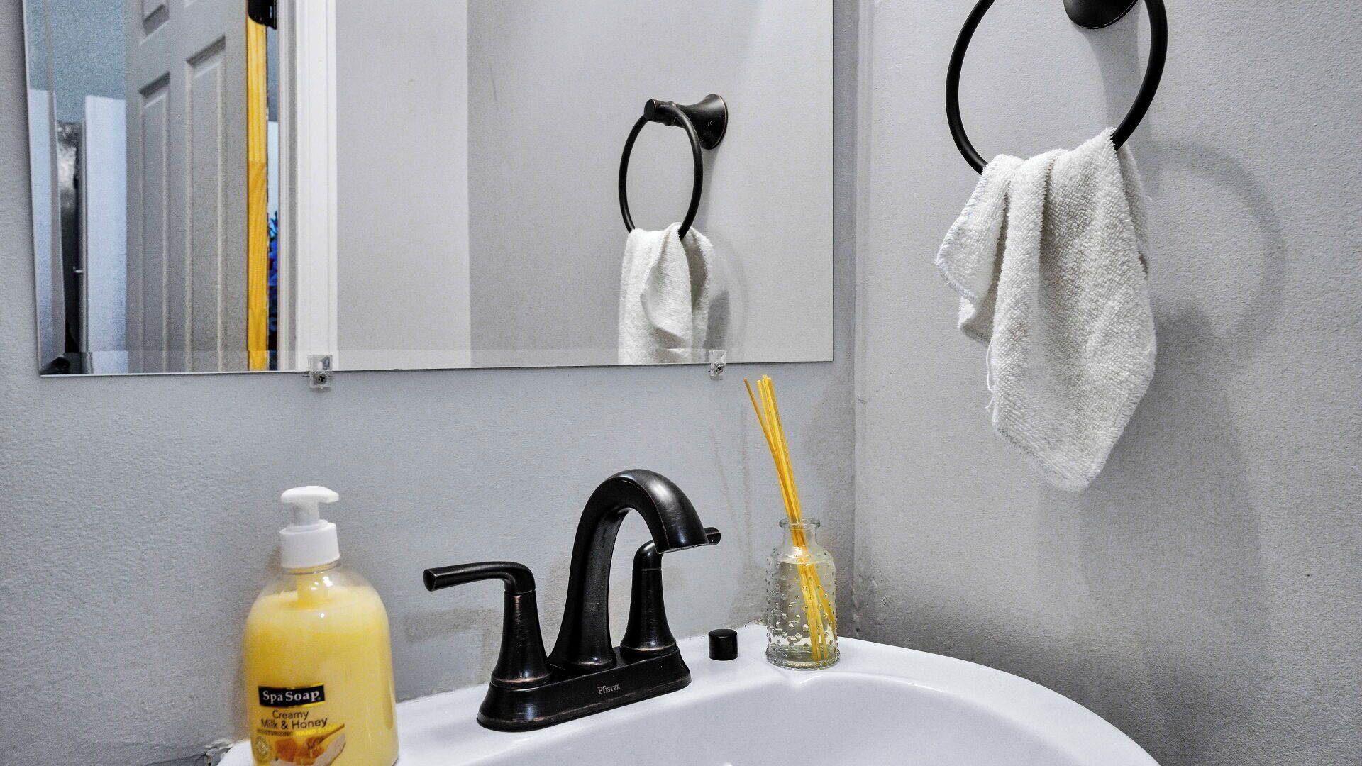 Une salle de bain avec un robinet noir, un flacon de savon, un verre contenant un diffuseur à bâtonnets et une serviette suspendue à un anneau.