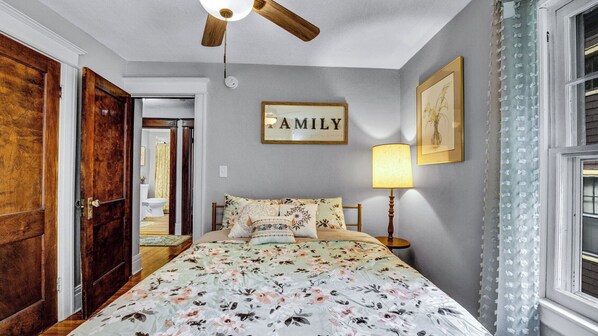 A bedroom with a wooden door, a ceiling fan, a bed with floral bedding, a bedside table with a lamp, and a framed 'FAMILY' sign on the wall. - Live in sophistication and elegance in your VRBO home (Rochester)