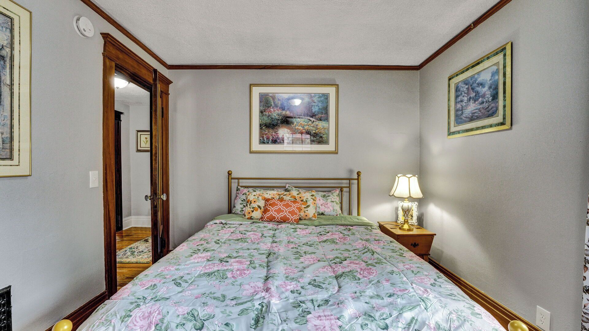Une chambre à coucher avec une couvre-lit à motifs floraux, une table de chevet avec une lampe et des tableaux encadrés accrochés aux murs.